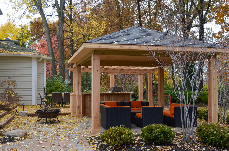 Wood pavilion with lounge seating, paver patio, shrubs, leaf-covered beds, and detached garage in fall.